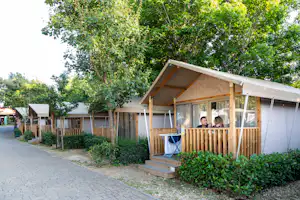 Holz- und Zeltbungalow mit Veranda und Sitzbank, am Wegesrand zwischen Büschen und Bäumen auf dem Campingplatz.
