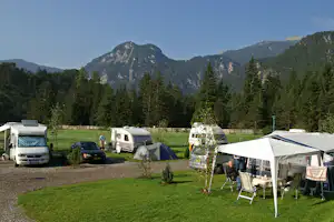 Campingplatz mit Zelten und Wohnmobilen auf einer Wiese, im Hintergrund hohe Berge und dichte Nadelwälder.