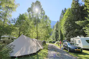 Zelt auf einem Campingplatz, daneben Wohnmobile und Autos, schattiger Weg durch Bäume und grünes Gras.