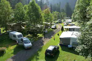Campingplatz mit Wohnwagen und Zelten, Autos am Weg, viel Grün und hohe Bäume im Hintergrund.