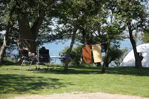 Campingplatz am See: Liegestühle und Tisch unter Bäumen, Wäscheleine mit Tüten, Zelt im Hintergrund.
