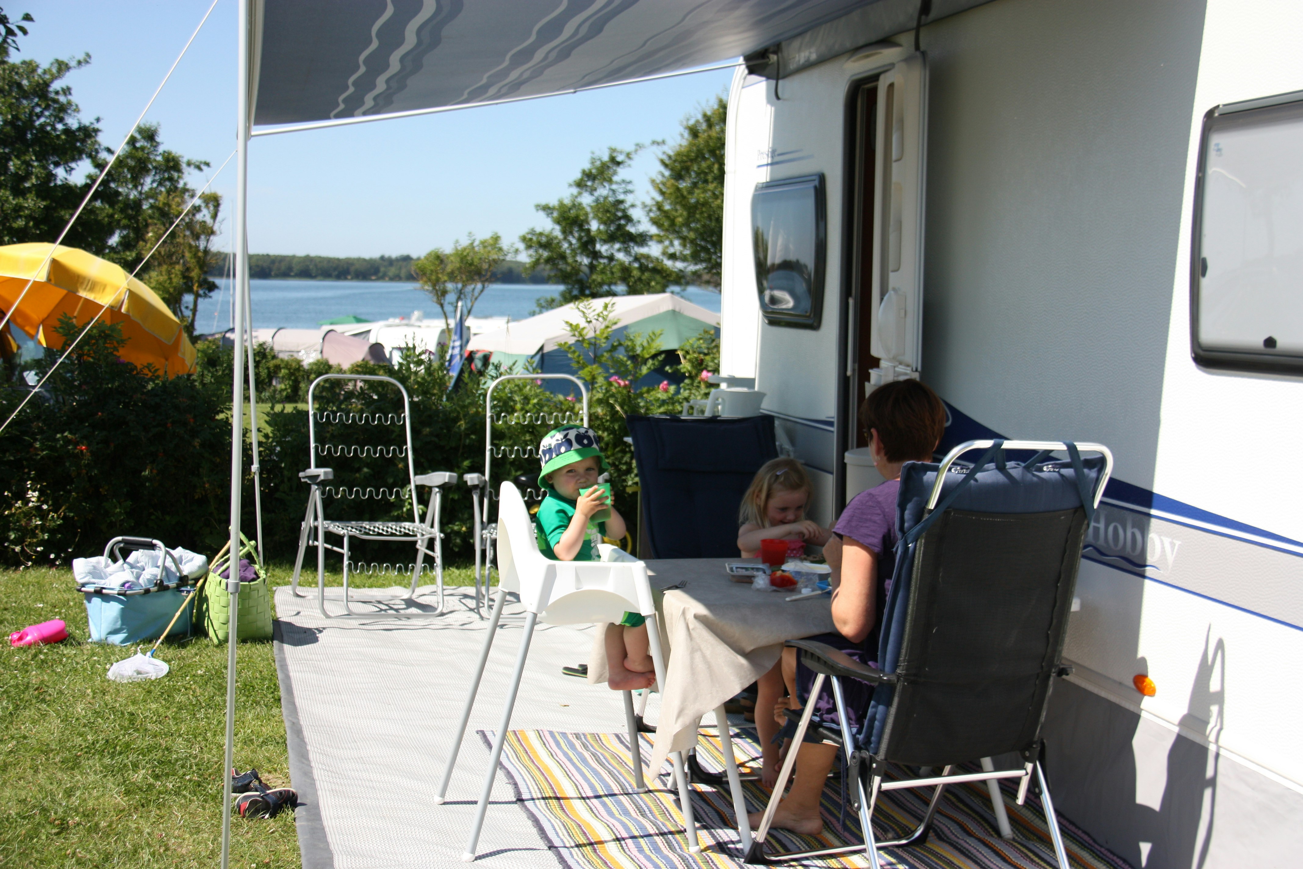 Wohnwagen-Terrasse mit Markise, Campingstühlen und Tisch, im Hintergrund Zelte und ein See zwischen Bäumen.