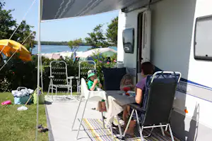 Wohnwagen-Terrasse mit Markise, Campingstühlen und Tisch, im Hintergrund Zelte und ein See zwischen Bäumen.