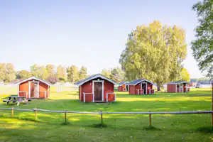 Rote Holz-Campinghäuschen mit Türen und Nummern auf einer grünen Wiese, umgeben von Bäumen und Zaun.