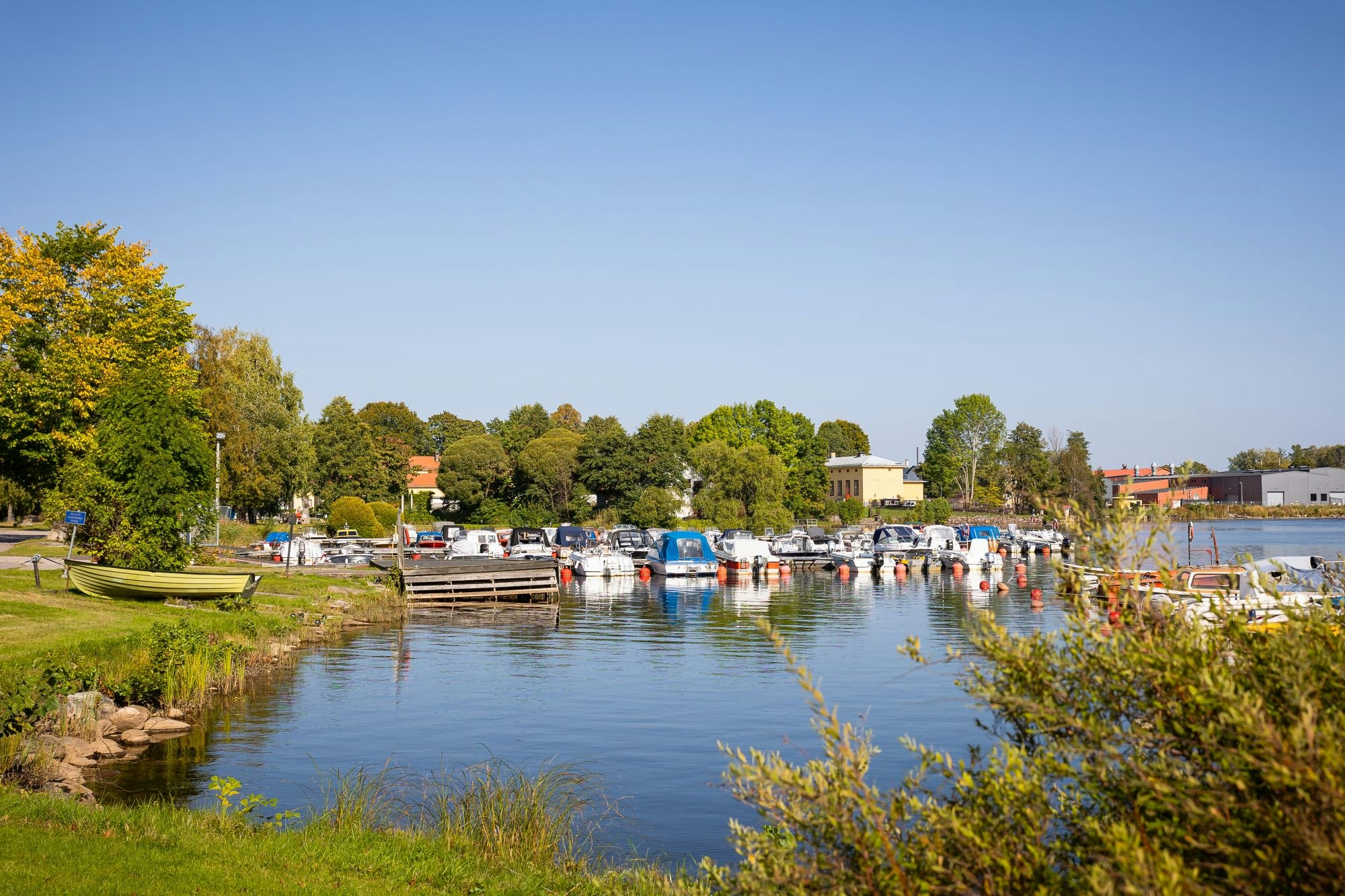 Hafen mit vielen Booten am Steg, Parkplatz mit Autos und Bäumen am Ufer eines ruhigen Gewässers.