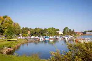 Hafen mit vielen Booten am Steg, Parkplatz mit Autos und Bäumen am Ufer eines ruhigen Gewässers.