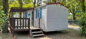 Kleines Mobilheim mit Veranda und Holztreppe im Grünen, umgeben von Bäumen und einem Holzgeländer.