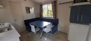 Wohnküche in einem Campingwagen mit Esstisch, zwei Stühlen, Küchenzeile und Fenster mit Vorhängen.