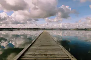 Holzsteg führt über den See, Spiegelung im Wasser, im Hintergrund Uferlinie und große Wolken am Himmel.
