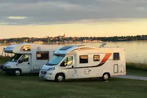 Wohnmobil und Kastenwagen am Ufer, Blick auf eine Stadt mit Kirchturm und Bäumen am Fluss bei Abendlicht.