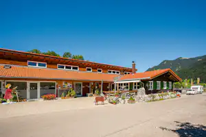Campinggebäude mit Holzfassade, Ziegeldach und Terrasse mit Sitzgelegenheiten, davor gepflasterter Platz und Blumenbeete.