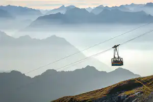 Seilbahn-Kabine an der Stütze über einem Bergpanorama mit Nebel und schroffen Gipfeln im Hintergrund.