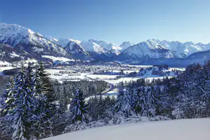 Winterliche Berglandschaft mit verschneiten Tannen, Tal mit Häusern und gewundenem Weg zwischen Wiesen und Feldern.