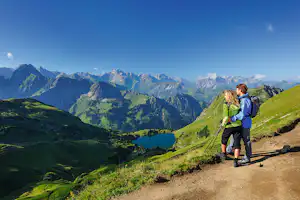 Wanderer auf einem Bergpfad mit Rucksack, Blick auf ein grünes Tal und die Alpenkette im Hintergrund.