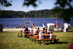 Camping am See mit Volleyballnetz, Picknicktischen und Zelten am Ufer, im Hintergrund Wald und Bojen im Wasser.