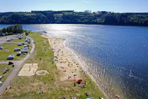 Campingplatz am See: Stellplätze mit Wohnmobilen und Autos, Grasstrand mit vielen Liegen und Blick auf bewaldete Hügel.