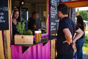 Mann am Tresen einer Campingbar, vor ihm ein Bierglas, im Hintergrund weitere Gäste und eine Tafel mit Kreide-Schrift.