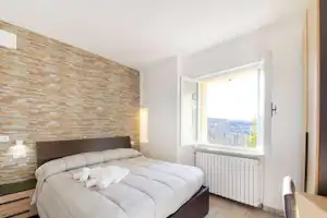 Schlafzimmer mit Doppelbett, weißer Wand und Fenster mit Blick ins Tal, Fliesenboden und Steinwand am Kopfteil.