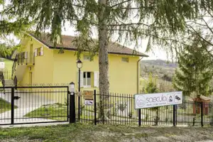 Gelbes Haus am Campingplatzzaun, mit Metallgitterzaun, Schildern und Bäumen im Vordergrund.