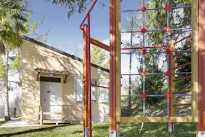 Wohnwagen mit Terrasse und Zaun, im Hintergrund Bäume und ein Spielgerüst aus Holz und Seilen.