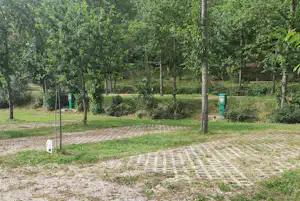 Campingplatz mit gepflastertem Weg, Rasenfläche und Baumreihe, im Hintergrund grüne Hecken und ein Sitzbereich.
