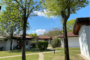 Campinghaus mit weißer Fassade, gepflastertem Weg und Rasenfläche, umgeben von Bäumen und Hecken im Garten.