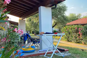 Terrasse am Campingplatz mit weißen Liegestühlen, blauen Aufblasartikeln und Blumen vor einem Garten mit Bäumen und Hausdach.