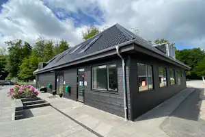 Ferienhaus auf dem Campingplatz mit Terrasse, gepflastertem Weg, Gartenpflanzen und großen Fenstern unter bewölktem Himmel.