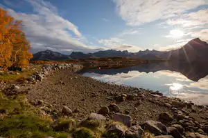 Seeufer mit Kiesstrand, ruhigem Wasser und Bergen in der Ferne, im Vordergrund Steine und Gras.