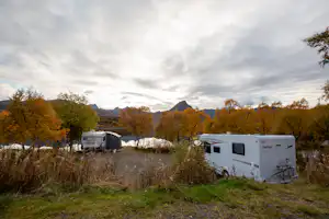 Wohnwagen auf einem Campingplatz am Waldrand, im Hintergrund Berge und herbstliche Bäume.