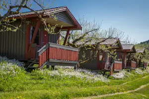 Rote Holz-Campinghütten mit Veranda und Treppe, umgeben von grünem Rasen, Blumen und Bäumen im Hintergrund.