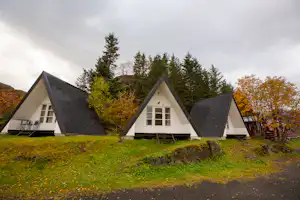 Drei weiße Ferienhäuser mit schwarzen Giebeldächern auf einer grünen Wiese, davor Wege und Sitzgelegenheiten, umgeben von Bäumen.