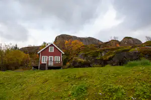 Rotes Ferienhaus mit Veranda auf einer grünen Wiese, umgeben von Bäumen und Bergen im Hintergrund.