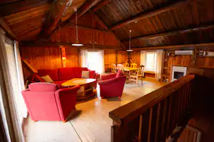Wohnzimmer in einem Holzhaus mit roter Sitzgruppe, Esstisch und Stühlen unter sichtbaren Holzbalken.
