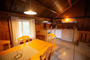 Innenraum eines Holzhauses mit Esstisch, gelben Stühlen und Küchenzeile mit Schränken und Herd.