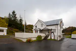 Weiße Campinghaus-Anlage mit Treppe, Eingang und Fahnenmast, daneben ein Weg und Bäume am Hang.