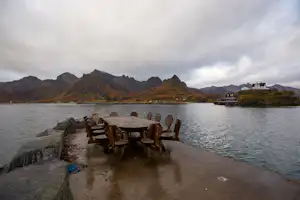 Picknicktisch auf einem Steg am See, im Hintergrund Berge und ein Ufer mit Gebäuden.