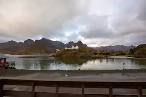 Blick vom Campingplatz auf einen See mit Uferpromenade, Steg und Bergen in der Ferne unter bewölktem Himmel.