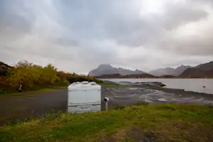Wohnwagen am Flussufer auf einem Campingplatz, Straße und Wiese im Vordergrund, Berge in der Ferne unter Wolken.