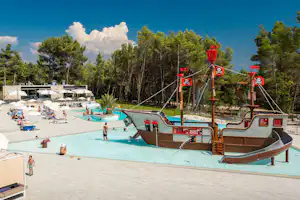 Schwimmbecken auf dem Campingplatz mit Spielboot, Liegen und Wegen, umgeben von Bäumen und einem Parkplatz.