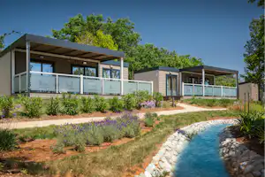 Moderne Camping-Bungalows mit Terrasse, Glasgeländer und gepflegtem Gartenweg, daneben ein kleiner Wasserlauf aus Steinen.