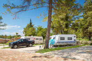 Stellplatz am Campingplatz mit Wohnwagen, Auto und gepflastertem Weg, umgeben von Bäumen und Grünflächen.