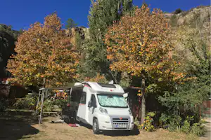 Weißes Wohnmobil auf dem Campingplatz, im Hintergrund Felsen und bewaldete Hänge, davor Bäume mit buntem Laub.