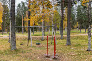 Spielplatz auf dem Campingplatz mit Schaukeln und rotem Reck im Herbstwald, umgeben von Bäumen und Rasen.