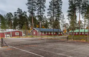 Tennisplatz mit Umzäunung und Asphaltfläche, dahinter rote Ferienhäuser und Bungalows im Wald mit hohen Bäumen.
