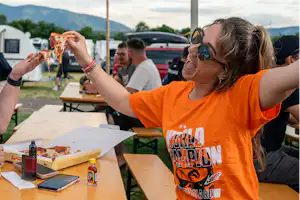Camping-Tisch unter einem großen Zelt, Personen beim Essen, Grillgut und Getränke auf dem Tisch, im Hintergrund Fahrzeuge.