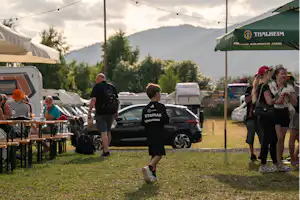 Campingplatz mit mehreren Zelten, Wohnmobil und Autos, Menschen am Tischbereich und Wimpelketten vor Bergen im Hintergrund.