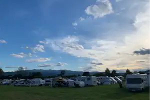 Campingplatz mit Wohnwagen auf einer grünen Wiese, im Hintergrund Berge und ein weiter Himmel mit Wolken.