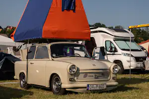Ein Wohnmobil mit aufgesetztem Zelt, daneben weitere Camper auf einem Campingplatz mit Rasen und Bäumen im Hintergrund.