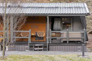 Camping-Villa mit Holzfassade, Terrasse und Markise, davor zwei Stühle und Tisch, im Hintergrund Bäume und Kiesweg.